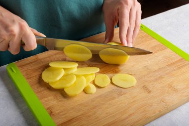 Hakiki kadın elleri mutfak masasında tahta kesme tahtasıyla patates kesiyor. Önlüklü kadın patates kesimi, yemek hazırlama, ev içi, ev hayatı, yaşam tarzı.