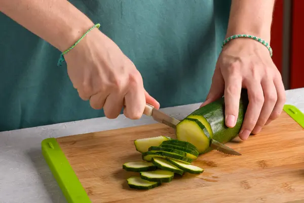 Authentic female hands cutting zucchini on wooden cutting board on kitchen table. Woman in apron cut zucchini for preparing food. Vegan and vegetarian recipe, cuisine. Domestic life, lifestyle