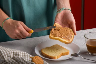 Hakiki kadın elleri, gri mutfak masasında beyaz ekmek ekmeğine fıstık ezmesi sürüyor, yakın plan. Tipik atıştırmalık yiyecekler, yemek yaşam tarzı, ev hayatı, Amerikan kahvaltısı hazırlama.