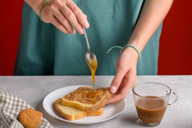 Kadınların elleri kahvaltıda yemek yapmak için fıstık ezmeli tostun üzerine kaşıkla bal koyuyor. Gri mutfak masasında, yakın plan. Fıstık ezmeli sandviçe damlayan bal.