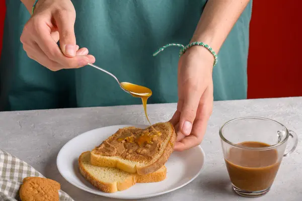 Womans hands puting honey with a spoon on a toast with peanut butter to make a sandwich for breakfast, at gray kitchen table, close-up. honey dripping from a spoon onto a peanut butter sandwich