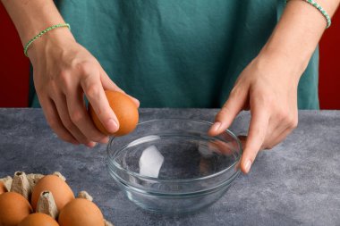 Bir kadının eli, gri mutfak masasındaki cam bir kaseye yumurta kırıyor. Yemek pişirme süreci, yaşam tarzı, yakın çekim.