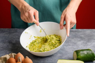 Çatalla karıştırılan kadın elleri çırpılmış yumurtalar ve gri mutfak masasındaki cam bir kasede rendelenmiş kabak. Kahvaltıda çırpılmış yumurta ve kabak pişirmek, hafif ve sağlıklı akşam yemeği pişirmek.