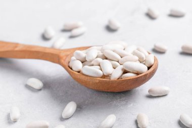 Gri bir masa yüzeyinde tahta bir kaşıkta beyaz fasulye. Akdeniz diyetlerinde popüler olan bu baklagiller, genellikle çorba ve güveçte kullanılan lif ve protein yönünden zengindir.