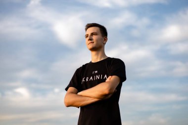 Bir adam mavi gökyüzünün arka planına karşı buğday tarlasında duruyor elleri önünde katlanmış bir şekilde Ukraynalı olduğu yazılı bir tişörtle.