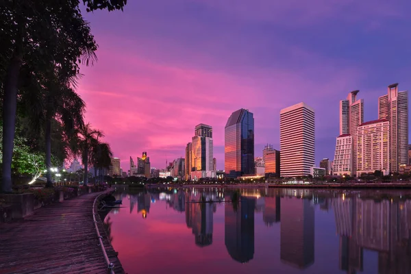 stock image Beautiful landscape with modern high-rise buildings reflecting in calm lake waters by park and boardwalk at colorful sunset. Benjakitti Park, Bangkok, Thailand.