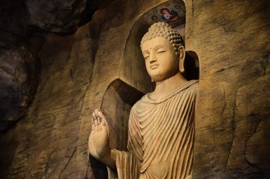 Kireçtaşı mağarasına oyulmuş ve ışıkla aydınlatılmış keşiş kıyafetleriyle ayakta duran Buda heykeli. Sağ el Adhay Mudra jesti. Din, gelenek, dua, meditasyon. 