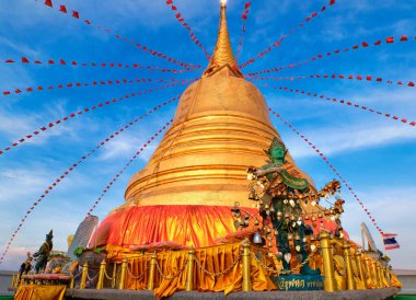 Statue of green-skinned Thai angel holding roll with message in Thai - translation: free from sorrow - stand in front of big golden stupa in sunlight and at blue sky with clouds in Buddhist temple. Decorations with bells, golden leaves, red flags. 