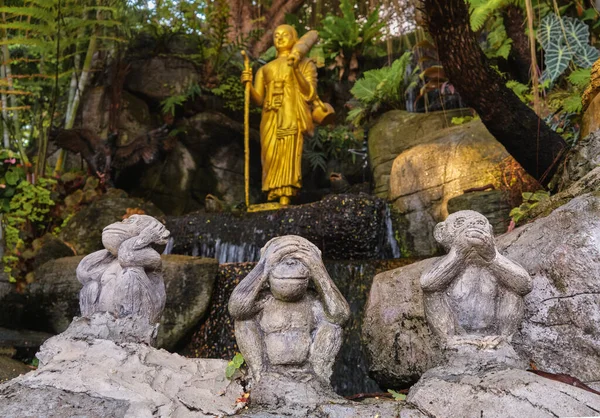 Buddhism symbols: gilded statue of Buddha as walking monk with Naga walking stick and umbrella and three wise monkeys statues sitting in foreground. Religion, diversity, inclusivity, prayer, worship.