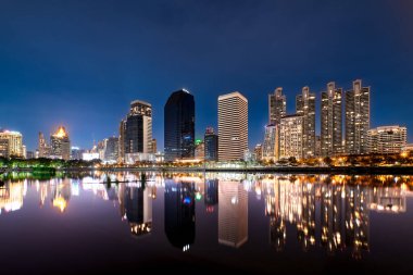 Beautiful landscape with modern high-rise buildings reflecting in calm lake waters at night. Benjakitti Park, Bangkok, Thailand.