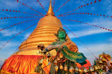 Statue of green-skinned Thai angel holding roll with message in Thai stand in front of big golden stupa in sunlight and at blue sky with clouds in Buddhist temple. Decorations with bells, golden leaves and red flags. Translation: free from sorrow