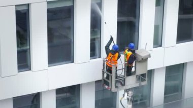 Güvenlik kemeri takan iki işçi, duvar, paneller ve ofis binasının ön cephesi önünde vinç beşiği ya da hava platformunda basınçlı temizleyici, silecek ve paspas kullanarak duruyor. Yakın çekim