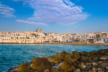 Geleneksel Yunan beyazlatılmış evleri, güneşli bir günde deniz kenarında, Yunanistan 'ın Paros Adası, Naoussa' da, tepede Tanrı 'nın Annesi' nin Dormition Kilisesi. Rıhtım ve tekneler limana demirlemiş, açık mavi gökyüzü, yaz