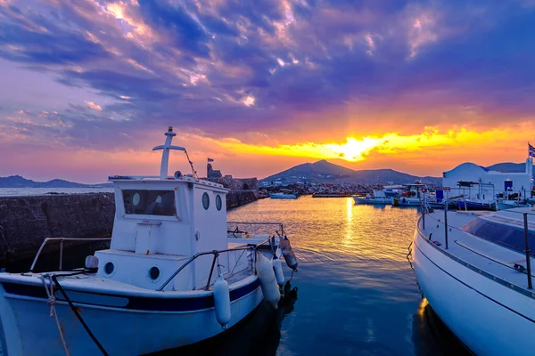 stock image Colorful sunset over quiet Greek fishing village and its harbour, old traditional boat moored by pier, sun rays and clouds in dramatic sky. Distant hills and hazy mountains, beautiful landscape or