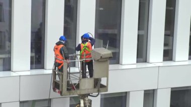 Güvenlik kemeri takan iki işçi, duvar, paneller ve ofis binasının ön cephesi önünde vinç beşiği ya da hava platformunda basınçlı temizleyici, silecek ve paspas kullanarak duruyor. El bilgisayarı