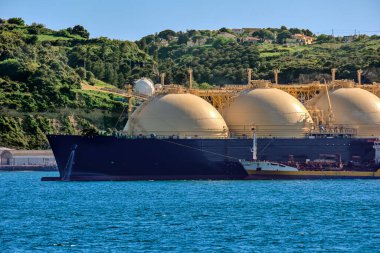 Güneşli bir günde endüstriyel rıhtım ve yeşil tepenin demirlediği sıvılaştırılmış doğal gaz tankeri. Alternatif gaz arzı, ticari taşımacılık, enerji krizi, yeniden gazlanma, çeşitlendirme, yakıt ve enerji