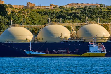 Güneşli bir günde endüstriyel rıhtım ve yeşil tepenin demirlediği sıvılaştırılmış doğal gaz tankeri. Alternatif gaz arzı, ticari taşımacılık, enerji krizi, yeniden gazlanma, çeşitlendirme, yakıt ve enerji