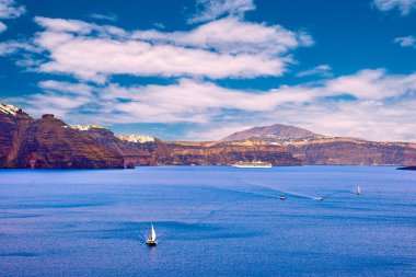 Santorini Adası Panorama, Yunanistan. Volkanik adanın engebeli uçurumları, güneşli bir gün, kaldera körfezi, mavi gökyüzü ve bulutlar, tepedeki Fira veya Thira kasabası, limandaki yelkenliler ve yatlar. Yaz gezisi