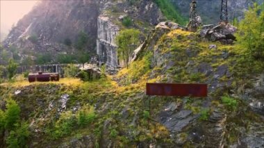 İtalya, Carrara, Toskana 'daki Apuan Alps dağlarındaki terk edilmiş mermer ocağının üzerinden havadan çekilen görüntü. İnsansız hava aracı yüksek ormanlık kayaların ve endüstriyel atıkların önünde parlak güneş ışığına karşı eğiliyor.