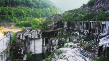 İHA 'nın hava görüntüsü yavaşça iniyor ve terk edilmiş mermer ocağının içinde dönüyor, Apuan Alps dağları, Carrara, İtalya. Siyah beyaz yaşlanmış, yıpranmış meşhur Carrara mermeri, yemyeşil yapraklar.