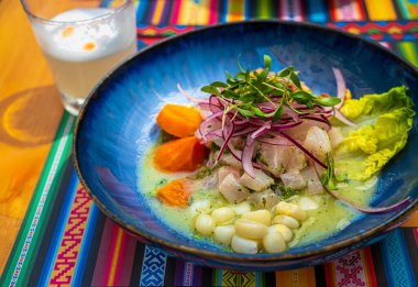 Geleneksel desenli mavi tabakta servis edilen taze ceviche ve Pisco Sour kokteyli. Seçici odaklanma, bulanık arkaplan. Çiğ balık, kırmızı soğan ve mısırdan yapılmış Peru usulü ceviche.