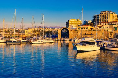 Yunanistan 'ın Girit adasının başkenti Heraklion limanında gün batımı. Şehir güvertesine demirlemiş balıkçı tekneleri, yatlar ve yelkenliler ve Arsenal ya da Neoria adlı eski Venedik liman harabeleri. Açık mavi gökyüzü