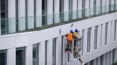 Güvenlik kemeri takan iki işçi ofis binasının duvarlarını, panellerini ve pencerelerini silecekler, paspaslar ile silecekler ile sileceklerin yüksekliğinde vinç beşiğinde ya da basınç makinesinin ardından hava platformunda duruyor. Çapraz