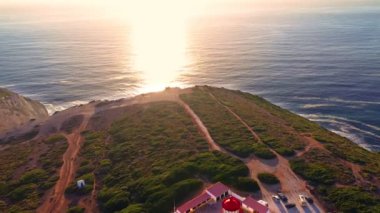 Günbatımında Cabo Espichel veya Cape Espichel 'deki deniz fenerinin insansız hava aracı görüntülerini geri çek. Atlantik Okyanusu kıyısı, Sesimbra, Portekiz. Uç, ufuktan, güneşten, parlaklıktan, yüksek kayalıklardan, Portekizce