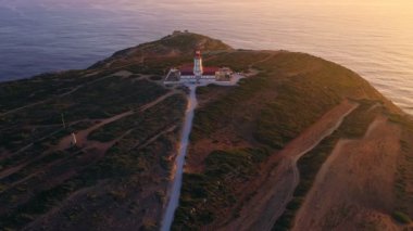Gün batımında Cabo Espichel ya da Cape Espichel 'deki deniz fenerinin insansız hava aracı görüntüsüne yaklaşıyoruz. Atlantik Okyanusu kıyısı, Sesimbra, Portekiz. Geniş görüş, yakınlaşma, çekilme, yüksek uçurumlar, Portekiz mimarisi