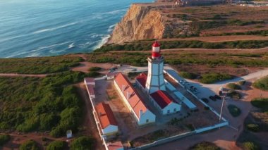 Gün batımında Cabo Espichel ya da Cape Espichel 'deki deniz fenerinin insansız hava aracı görüntüsü. Atlantik Okyanusu kıyısı, Sesimbra, Portekiz. Yörünge, parallaks, yüksek uçurumlar, Portekiz mimarisi, günbatımı yazı