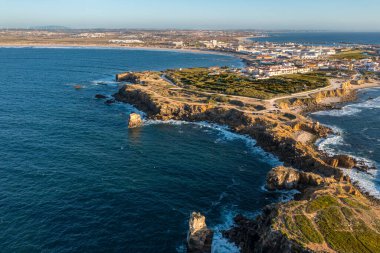 Peniche, Portekiz 'in kayalık kıyılarında mavi dalgaların havadan görünüşü bozuluyor. Yaz günbatımı sisi, biraz yeşillik ve kayalık uçurumlar, yarımada ve kayalar, balıkçı kasabası, ufuk.. 