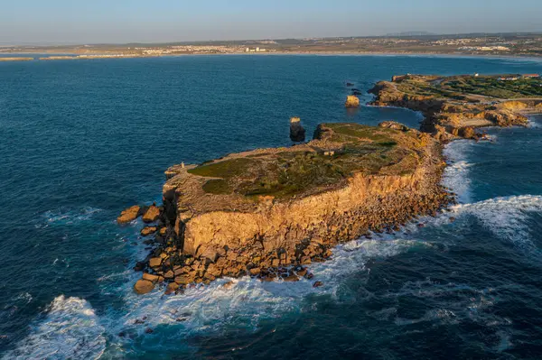 Aerial image of high rocky headland and peninsula. Drone shot of sea blue waves beat and splash in summer sunset haze, little foliage and rocky cliffs, establishing or static shot, Peniche, Portugal.