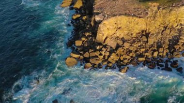 Kayalık kıyılarda okyanus dalgalarının görüntüsünü oluşturan drone kırılıyor. Mavi deniz dalgaları yaz günbatımında sisleri, yüksek kayalıkları ve büyük taşları vurur ve sıçratır. İnsanlar burnun kenarındaki manzaranın tadını çıkarırlar.