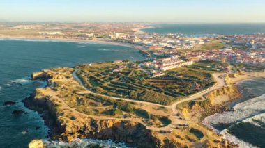 Portekiz 'in Peniche adası ve kasabasının insansız hava aracı görüntüsü, akşam güneşinde okyanus dalgaları kayalık kayalıklara vurur. Çekim ayarlanıyor, yavaşça iniyor ya da yukarıdaki güzel balıkçı kasabasının havadan görüntüsü aşağı uçuyor.