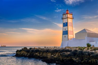 Deniz kıyısında renkli bir gün batımı ve Portekiz, Cascais 'de Santa Marta deniz feneri. Renkli gökyüzü, bulutlar, güneş ışığı, alçak güneş, sakin okyanus suları, yerel simge, güvenlik ve seyrüsefer kulesi, kayalık kıyı. 