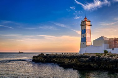 Deniz kıyısında dramatik bir gün batımı ve Portekiz, Cascais 'de Santa Marta deniz feneri. Renkli gökyüzü, bulutlar, güneş ışığı, alçak güneş, sakin okyanus suları, yerel simge, güvenlik ve seyrüsefer kulesi, kayalık kıyı. 