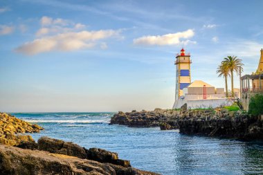 Deniz kıyısında güneşli bir gün ve Cascais, Portekiz 'de Santa Marta deniz feneri. Renkli gökyüzü, bulutlar, güneş ışığı, palmiye ağacı, sakin okyanus suları, yerel dönüm noktası, güvenlik ve seyrüsefer kulesi, kayalık kıyı