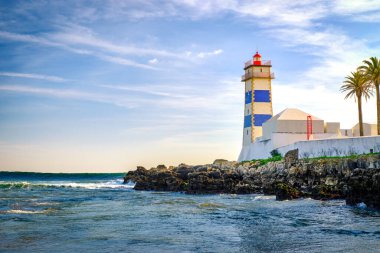 Güzel bir akşam, deniz gelgit dalgaları, kayalık kıyılar ve Cascais, Portekiz 'deki Santa Marta deniz feneri. Mavi gökyüzü, bulutlar, güneş ışığı, alçak güneş, sakin okyanus suları, yerel dönüm noktası, güvenlik ve seyrüsefer kulesi