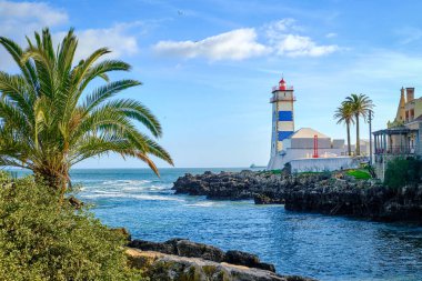 Deniz kıyısında güneşli güzel bir gün ve Cascais, Portekiz 'de Santa Marta deniz feneri. Açık mavi gökyüzü, bulutlar, güneş ışığı, palmiye ağaçları, sakin okyanus suları, yerel dönüm noktası, güvenlik ve seyrüsefer kulesi, kayalık
