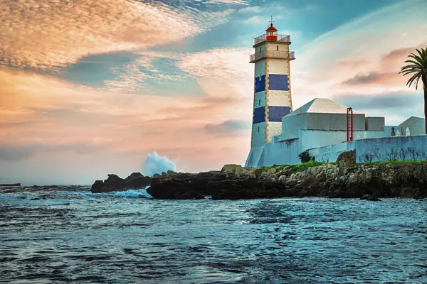 Deniz kıyısında dramatik bir gün batımı ve Portekiz, Cascais 'de Santa Marta deniz feneri. Renkli gökyüzü, bulutlar, güneş ışığı, alçak güneş, sakin okyanus suları, yerel simge, güvenlik ve seyrüsefer kulesi, kayalık