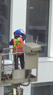 Güvenlik kemeri takan iki işçi, duvar, paneller ve ofis binasının ön cephesi önünde vinç beşiği ya da hava platformunda basınçlı temizleyici, silecek ve paspas kullanarak duruyor. El bilgisayarı
