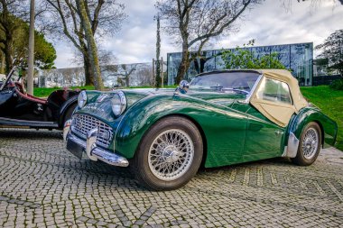 Lisbon, Portugal - Jan 20, 2024: Classic green Triumph TR3A soft top drophead car cobblestone driveways by another vintage vehicle on sunny spring day. clipart