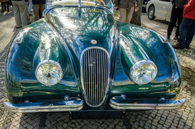 Lisbon, Portugal - Jan 20, 2024: Front view of vintage dark green Jaguar XK120 drophead coupe car parked on cobblestone pavement road. Two-seater roadster, sports car, convertible clipart