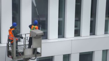 Güvenlik kemeri takan iki işçi, duvar, paneller ve ofis binasının ön cephesi önünde vinç beşiği ya da hava platformunda basınçlı temizleyici, silecek ve paspas kullanarak duruyor. Kopyalama alanı