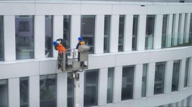 Güvenlik kemeri takan iki işçi, duvar, paneller ve ofis binasının ön cephesi önünde vinç beşiği ya da hava platformunda basınçlı temizleyici, silecek ve paspas kullanarak duruyor. Gün