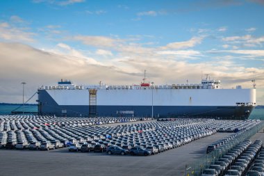 Large vehicle carrier ship known as RO-RO at port on sunny day, large parking area filled with cars. Blue sky and colorful clouds, vehicle transportation. clipart