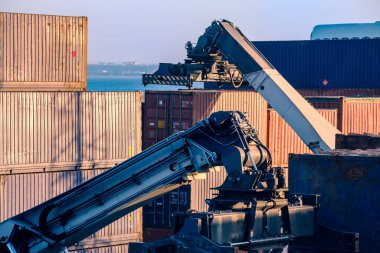 Reach stacker arms and spreaders maneuver while grabbing and carrying cargo containers in sea port shipping terminal on sunny day, clear blue sky.  clipart