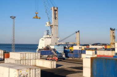 Ticari limandaki deniz lojistik ve ulaşım sahneleri çeşitli renklerde istiflenmiş nakliye konteynerleri, kargo işleme ekipmanları ve açık mavi gökyüzü arka planına karşı gemi vinçlerini gösteriyor.
