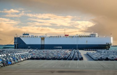 RO-RO olarak bilinen büyük bir araç ya da taşıyıcı gemi güneşli bir günde, limanda, içi araba dolu büyük bir otoparkta yuvarlanıyor. Mavi gökyüzü ve renkli bulutlar, araç ulaşımı.