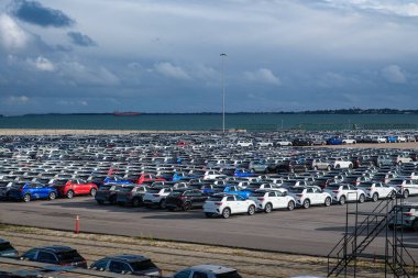 Liman kargo terminalindeki geniş bir otoparkın havadan görüntüsü. Sıralar dolusu yeni araç boşluğu doldurarak renkli bir mozaik oluşturuyor. Arabalar yakındaki Ro-Ro gemileriyle küresel varış noktalarına sevkiyatı bekliyor. Ölçekle